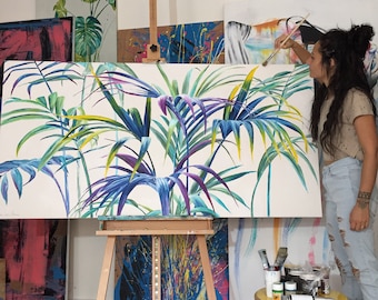 May include: A woman paints a large canvas with a tropical leaf design. The canvas is on an easel and the leaves are painted in shades of blue, green, and purple.