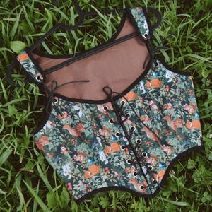 May include: A black and green corset with a woodland print featuring small animals and plants. The corset has a brown lining and black lace-up closure in the front.