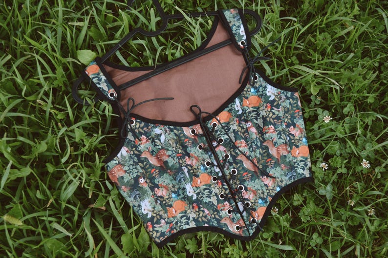 May include: A black and green corset with a woodland print featuring small animals and plants. The corset has a brown lining and black lace-up closure in the front.