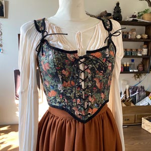 May include: A black corset with a forest print featuring owls, squirrels, and deer. The corset has black lacing and is worn over a white blouse and a brown skirt.