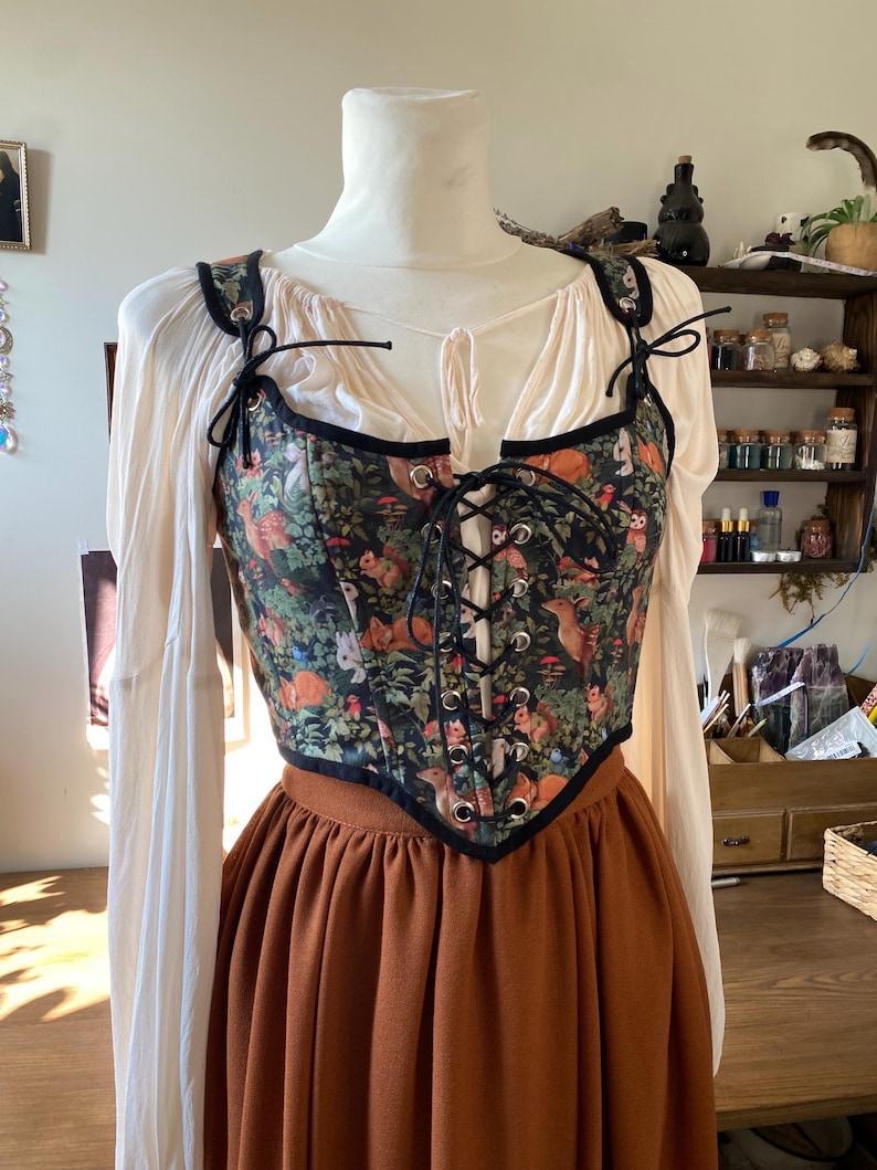 May include: A black corset with a forest print featuring owls, squirrels, and deer. The corset has black lacing and is worn over a white blouse and a brown skirt.