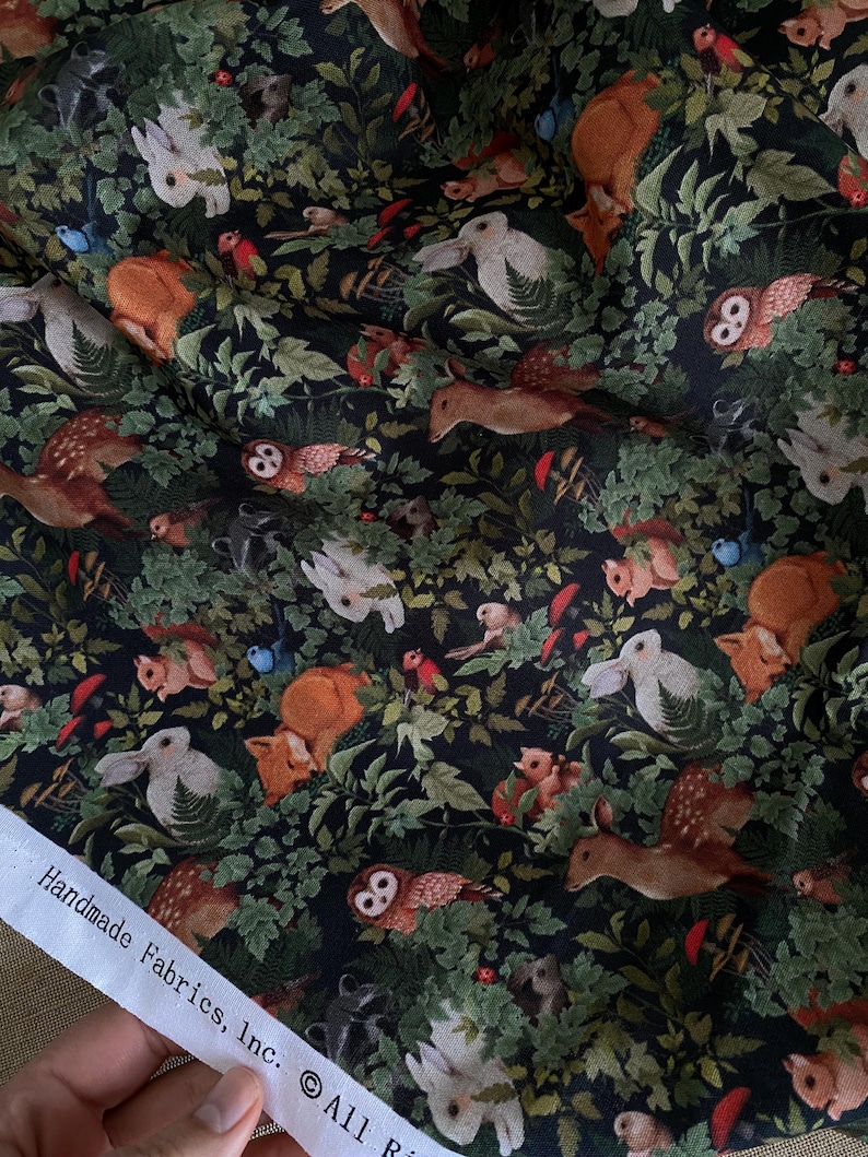 May include: Black fabric with a repeating pattern of white rabbits, brown deer, owls, squirrels, mushrooms, and red birds in a forest setting. The fabric has the text "Handmade Fabrics, Inc. &copy; All Ri" printed on the bottom edge.