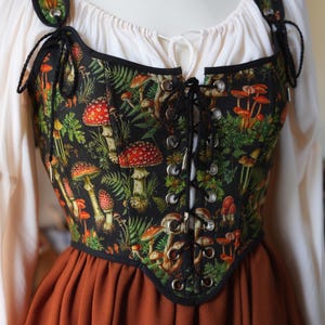 May include: A black corset with a colorful mushroom and fern pattern. The corset has a lace-up front and black trim. The straps are also patterned and attached with black cords. The corset is worn over a white blouse and a rust-colored skirt.