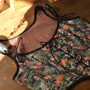 May include: A black and green corset with a woodland print featuring small animals and plants. The corset has a lace-up front and a black trim.