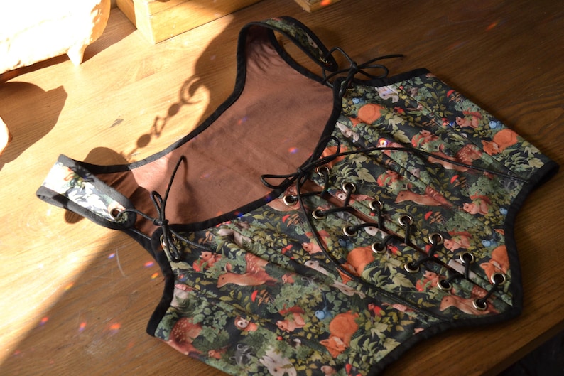 May include: A black and green corset with a woodland print featuring small animals and plants. The corset has a lace-up front and a black trim.