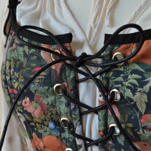 May include: A black corset with a forest print featuring animals and plants. The corset has black laces and silver eyelets.