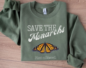 Könnte beinhalten: Ein olivgrünes Sweatshirt mit Rundhalsausschnitt, das den weißen Schriftzug "SAVE THE Monarchs" über einer Grafik eines Monarchfalters mit orangefarbenen, schwarzen und weißen Flügeln sowie den weißen Text "Plant milkweed." darunter zeigt.