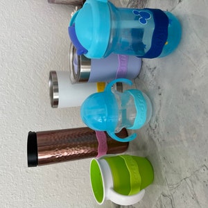 K&ouml;nnte beinhalten: Eine Sammlung von sechs verschiedenen wiederverwendbaren Wasserflaschen, darunter ein Edelstahlbecher, ein Kupferbecher, eine durchsichtige Plastikflasche mit blauem Deckel und blau-wei&szlig;em Strohhalm, eine blaue Plastikflasche mit blauem Deckel und blau-wei&szlig;em Strohhalm, eine durchsichtige Plastikflasche mit blauem Deckel und blau-wei&szlig;em Strohhalm und eine gr&uuml;ne Plastikflasche mit wei&szlig;em Deckel und wei&szlig;em Griff.