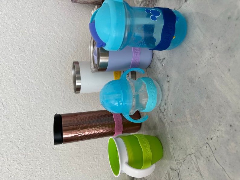K&ouml;nnte beinhalten: Eine Sammlung von sechs verschiedenen wiederverwendbaren Wasserflaschen, darunter ein Edelstahlbecher, ein Kupferbecher, eine durchsichtige Plastikflasche mit blauem Deckel und blau-wei&szlig;em Strohhalm, eine blaue Plastikflasche mit blauem Deckel und blau-wei&szlig;em Strohhalm, eine durchsichtige Plastikflasche mit blauem Deckel und blau-wei&szlig;em Strohhalm und eine gr&uuml;ne Plastikflasche mit wei&szlig;em Deckel und wei&szlig;em Griff.
