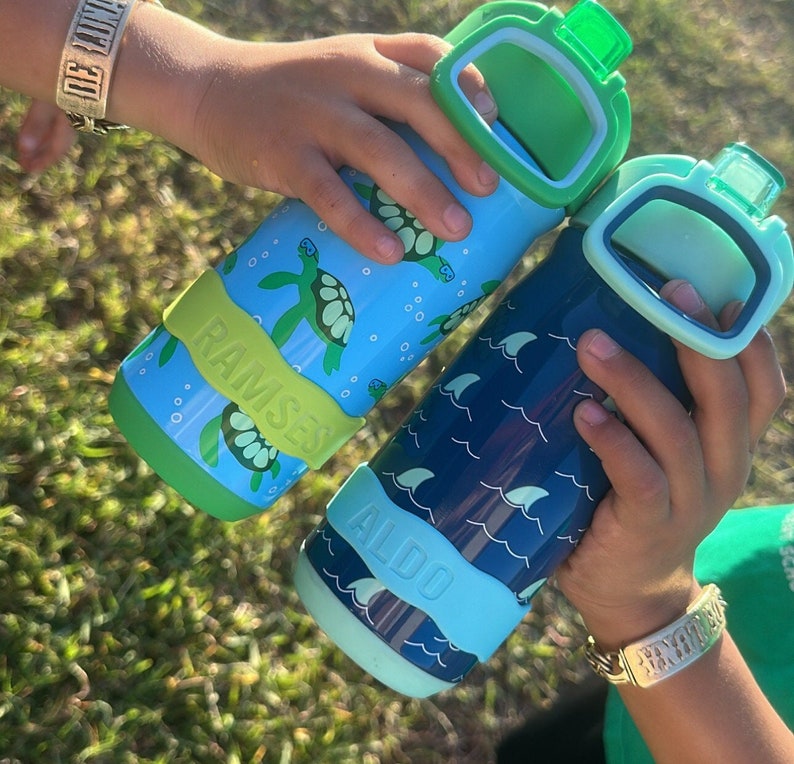 K&ouml;nnte beinhalten: Zwei wiederverwendbare Wasserflaschen mit bunten Designs. Eine Flasche ist blau mit einem Wellenmuster und dem Text "ALDO". Die andere Flasche ist blau und gr&uuml;n mit einem Schildkr&ouml;tenmuster und dem Text "RAMSES".