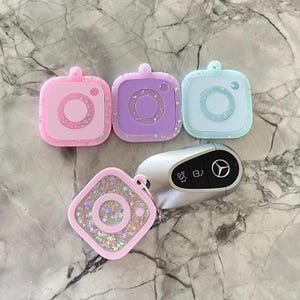 May include: A collection of colorful, square-shaped keychains in pink, purple, and blue, each featuring a camera design with a central circle and a small lens detail. The keychains are adorned with glitter and a silver key fob.