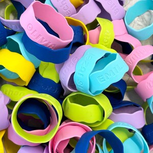 K&ouml;nnte beinhalten: Eine Sammlung bunter Silikon-Armb&auml;nder mit verschiedenen Namen, darunter "Olivia", "Emilie", "Is Izea", "Rebecca", "Amelia" und "Tilda".
