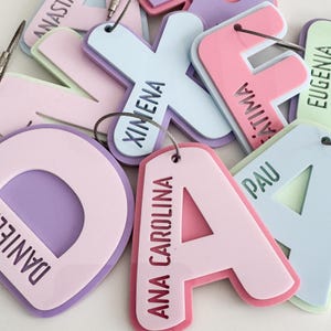 May include: A collection of colorful luggage tags with names, including "ANA CAROLINA", "XIMENA", and "ATIMA". The tags are in various pastel colors, such as pink, purple, and light blue, with silver-colored metal loops.