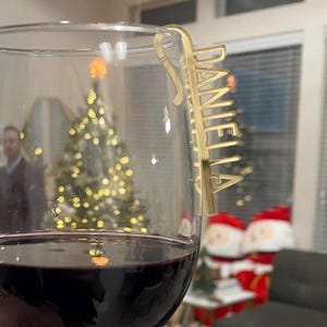 Könnte beinhalten: Ein goldfarbener Acryl-Weinglas-Anhänger mit dem eingravierten Namen "Daniella". Der Anhänger hängt am Rand eines Weinglases, das mit Rotwein gefüllt ist. Im Hintergrund ist ein Weihnachtsbaum mit Lichtern verschwommen.