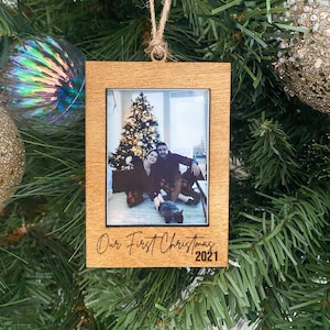 May include: A wooden Christmas ornament with a photo of a couple in front of a Christmas tree. The ornament is engraved with the text "Our First Christmas 2021".
