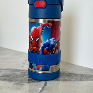 K&ouml;nnte beinhalten: Eine blaue Edelstahl-Trinkflasche mit rotem Deckel und einem Spiderman-Motiv. Die Flasche hat einen blauen Silikon-Gurt mit dem Namen "PEDRO" darauf.