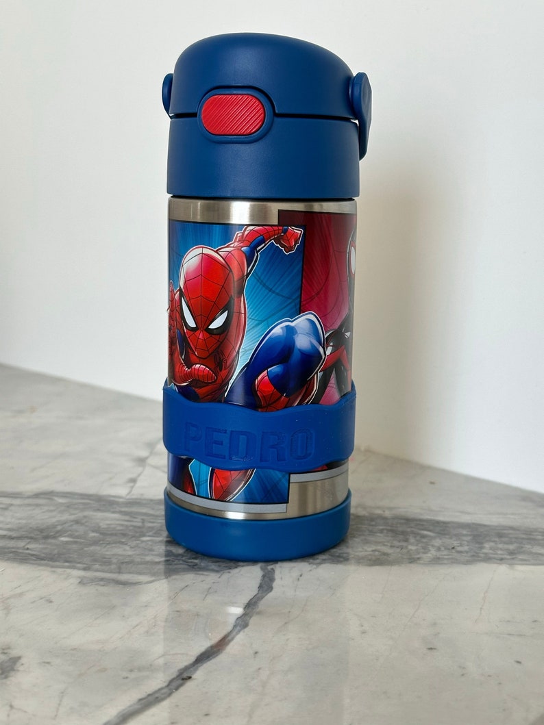K&ouml;nnte beinhalten: Eine blaue Edelstahl-Trinkflasche mit rotem Deckel und einem Spiderman-Motiv. Die Flasche hat einen blauen Silikon-Gurt mit dem Namen "PEDRO" darauf.