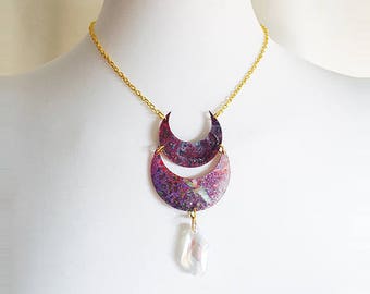 May include: A gold chain necklace with a double crescent moon pendant. The pendant is made of metal with a purple and red marbled finish. A white iridescent crystal dangles from the bottom of the pendant.