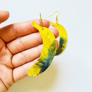 Puede incluir: Un par de pendientes amarillos en forma de plátano con detalles en negro y dorado. Los pendientes están hechos de resina y tienen un cierre de gancho dorado.