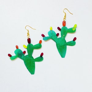 May include: A pair of green cactus earrings with red and yellow accents. The earrings are made of resin and have a gold hook.