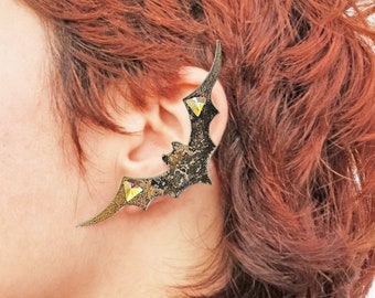May include: A gold-toned bat ear cuff with three clear rhinestones. The cuff is designed to wrap around the ear and has a textured finish.