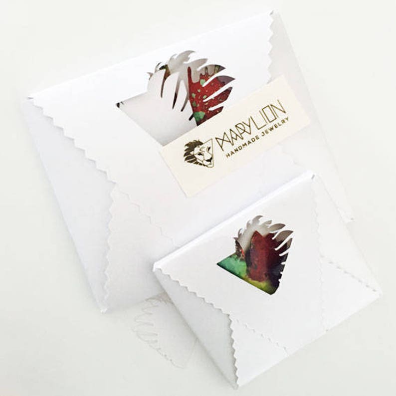 K&ouml;nnte beinhalten: Zwei wei&szlig;e Umschl&auml;ge mit gewellten Kanten. Der obere Umschlag hat ein goldenes Logo mit der Aufschrift "Mary Lion Handmade Jewelry". Der untere Umschlag hat ein ausgeschnittenes Design mit einem bunten Blumenmuster.