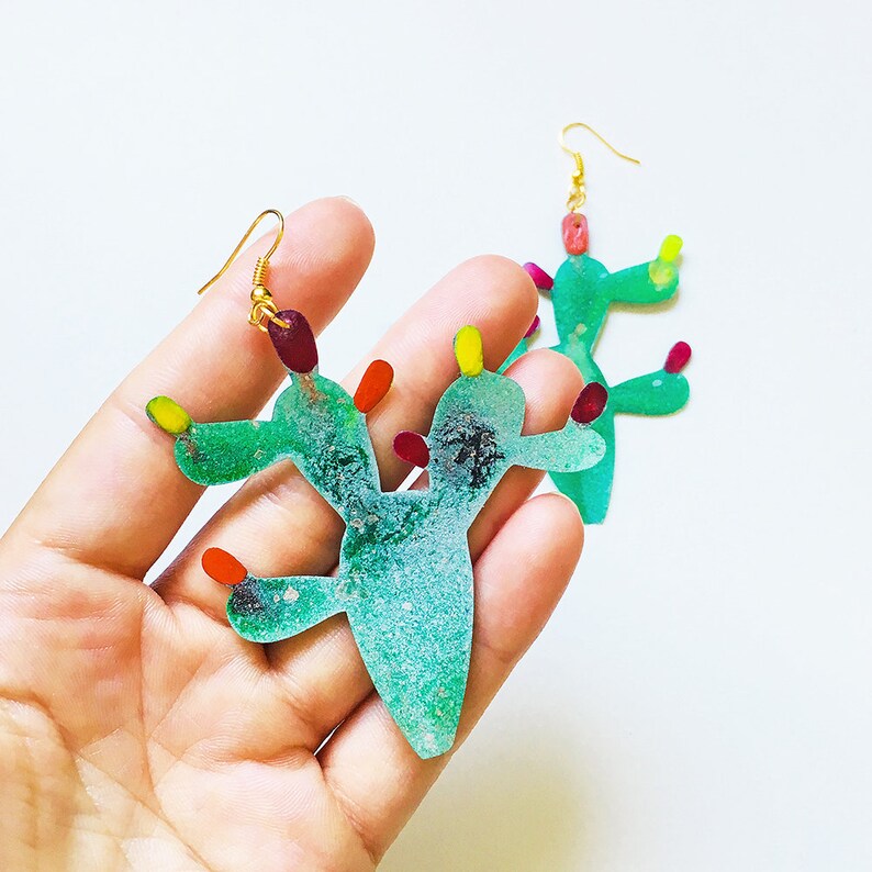 May include: A pair of green cactus earrings with colorful accents. The earrings are made of metal and have a textured finish.