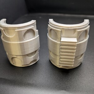 Puede incluir: Dos piezas de pl&aacute;stico gris impresas en 3D, cada una con forma de semicilindro con una peque&ntilde;a abertura rectangular en un lado. Las piezas parecen formar parte de una estructura m&aacute;s grande.