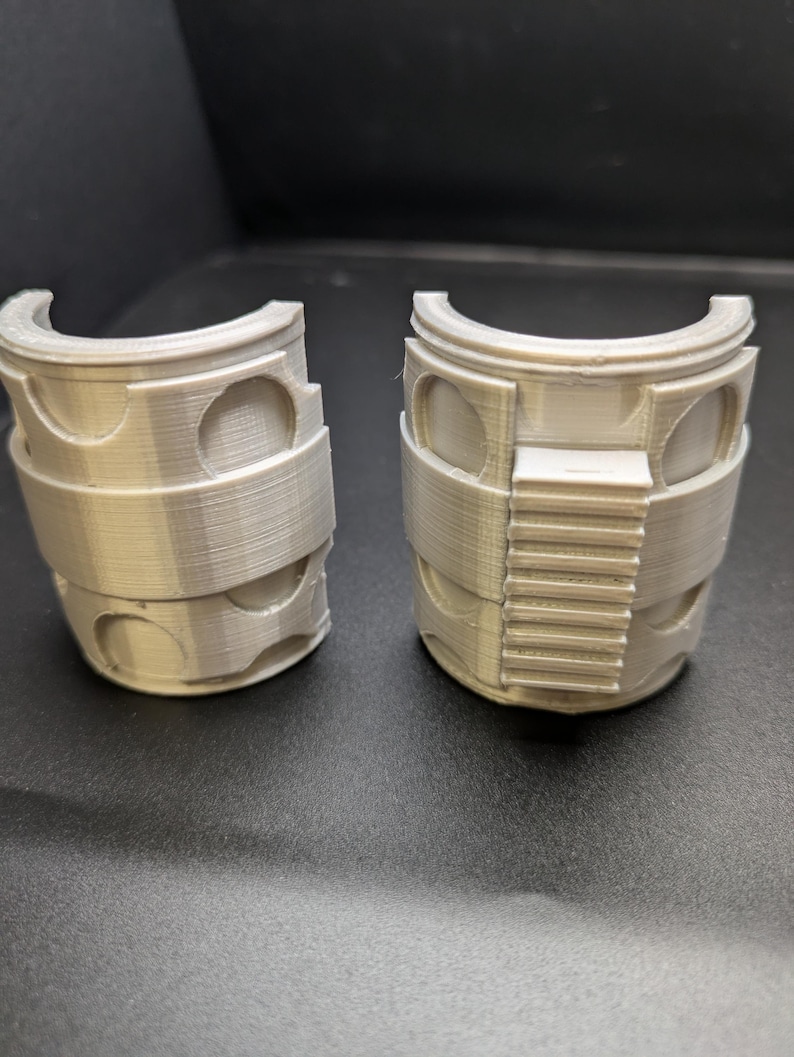 Puede incluir: Dos piezas de pl&aacute;stico gris impresas en 3D, cada una con forma de semicilindro con una peque&ntilde;a abertura rectangular en un lado. Las piezas parecen formar parte de una estructura m&aacute;s grande.