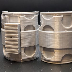 May include: A pair of gray 3D printed plastic cuffs with a textured surface and a series of circular cutouts. The cuffs are connected by a flat, textured band.