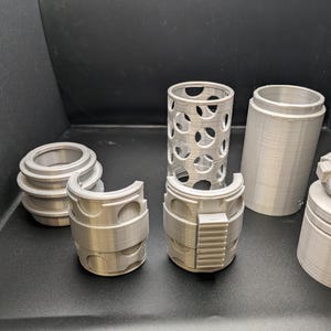 Puede incluir: Un conjunto de seis piezas de pl&aacute;stico de color plateado impresas en 3D para un modelo. Las piezas incluyen un cilindro con un patr&oacute;n de rejilla, un cilindro con una superficie lisa, un anillo con una superficie lisa, un anillo con un patr&oacute;n de rejilla, un anillo con una superficie lisa y un anillo con una superficie lisa y un dise&ntilde;o rectangular en relieve.