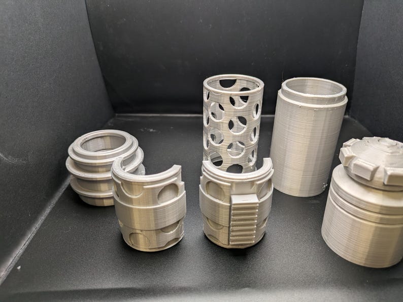 Puede incluir: Un conjunto de seis piezas de pl&aacute;stico de color plateado impresas en 3D para un modelo. Las piezas incluyen un cilindro con un patr&oacute;n de rejilla, un cilindro con una superficie lisa, un anillo con una superficie lisa, un anillo con un patr&oacute;n de rejilla, un anillo con una superficie lisa y un anillo con una superficie lisa y un dise&ntilde;o rectangular en relieve.