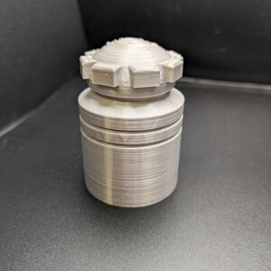 Puede incluir: Un tap&oacute;n de botella de color plateado, impreso en 3D, con un dise&ntilde;o texturizado y estriado. El tap&oacute;n tiene una parte superior circular con un patr&oacute;n de engranaje.