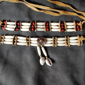 Peut inclure: Deux colliers en cuir et en os avec des perles rouges, noires et blanches. Le collier du bas a une pièce métallique décorative au centre et deux pendentifs en coquille.
