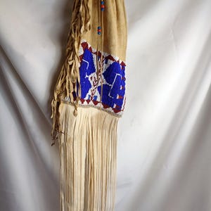 May include: A beaded Native American pouch with fringe. The pouch is made of beige leather and is decorated with blue, red, and white beads. The fringe is made of leather and is about 30 cm long.