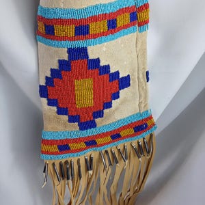 Op de afbeelding: Beige suède etui met ingewikkeld kralenwerk in rood, blauw en geel. Het ontwerp heeft geometrische patronen en een franje van bruine leren stroken. Metalen accenten zijn zichtbaar.