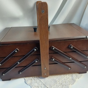 Könnte beinhalten: Eine Vintage-Nähkiste aus dunkelbraunem Holz. Sie hat einen Klappdeckel, einen Holzgriff und abgewinkelte Fächer. Der Griff ist aus hellem Holz gefertigt. Die Kiste steht auf einer weißen Spitzendecke.