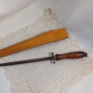 May include: A vintage knife sharpener with a wooden handle and a metal rod, alongside a tan leather sheath. The sharpener is designed for maintaining the sharpness of knives. The items are displayed on a white lace doily.
