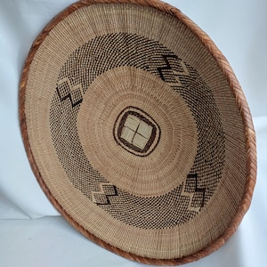 Könnte beinhalten: Ein großer, runder, geflochtener Korb mit braunem Rand und einem geometrischen Muster in Braun und Weiß. Der Korb ist aus natürlichen Materialien gefertigt und hat ein traditionelles Design.