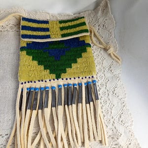 Könnte beinhalten: Kleine, rechteckige Perlenbeutel mit Kordelzug. Die Tasche hat einen gelben Hintergrund mit blauen und grünen geometrischen Mustern. Der untere Rand hat cremefarbene Fransen mit blauen Akzenten. Die Tasche wird auf einer cremefarbenen Spitzendecke präsentiert.