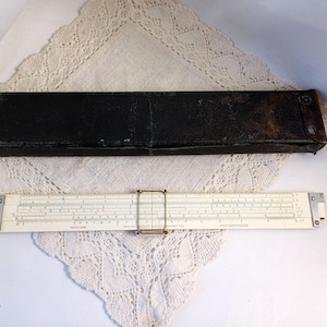 Könnte beinhalten: Ein antiker Rechenschieber mit cremefarbenem Gehäuse und schwarzem Etui. Der Rechenschieber weist detaillierte Markierungen und einen Metallrahmen auf. Das Etui ist schwarz und rechteckig und weist Gebrauchsspuren auf. Der Rechenschieber ist etwa 30 cm lang.