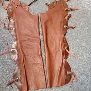 Op de afbeelding: Bruine leren chaps met decoratieve geschulpte randen en zilverkleurige metalen accenten. De chaps hebben verstelbare riemen en banden. Een klassiek Western-item.