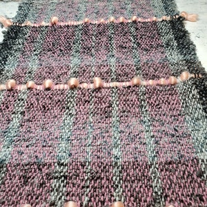 Peut inclure: Une couverture tissée avec un motif à carreaux rose et gris. La couverture a une bordure à franges avec des pompons roses.