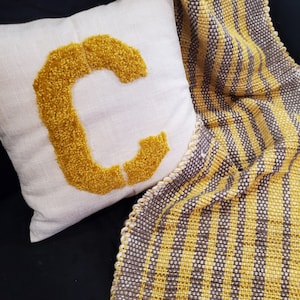 May include: A white throw pillow with a large, yellow, fluffy letter "C" embroidered on it. The pillow is sitting on a black couch with a yellow and gray striped throw blanket with fringe.