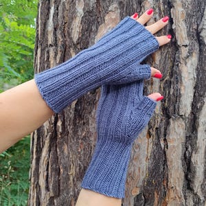 May include: A pair of blue fingerless gloves made of a ribbed knit fabric. The gloves are a muted blue color and appear to be handmade. The gloves are displayed against a tree trunk background.