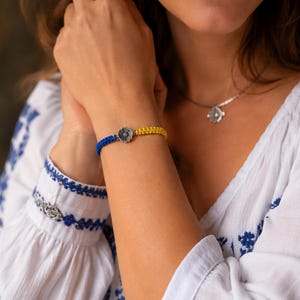 May include: A braided cord bracelet featuring a vibrant blue section and a bright yellow section, connected by a central silver-toned metal flower charm with intricate petal detailing and a textured center, creating a distinct two-tone appearance.