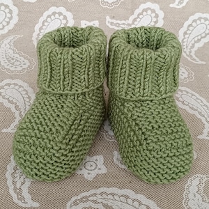 May include: A pair of olive green knitted baby booties. The booties have a ribbed cuff and a textured knit pattern. The booties are designed to keep a baby's feet warm and cozy. The booties are made of soft yarn.