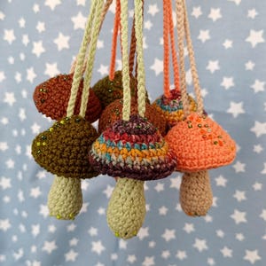 May include: A collection of crocheted mushroom ornaments with colorful yarn and beaded accents. The mushrooms are hanging from strings and have different colors and patterns.