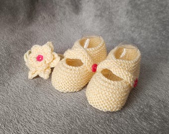 May include: A pair of pale yellow knit booties with a textured garter stitch pattern, each featuring a single strap secured by a small pink button, accompanied by a matching pale yellow crocheted flower accessory with a pink center button.