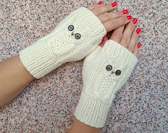 May include: A pair of cream-colored fingerless gloves featuring a cable-knit owl design on the back of each hand. The owl motifs are accented with two small, dark circular buttons serving as eyes, integrated into the ribbed knit fabric.